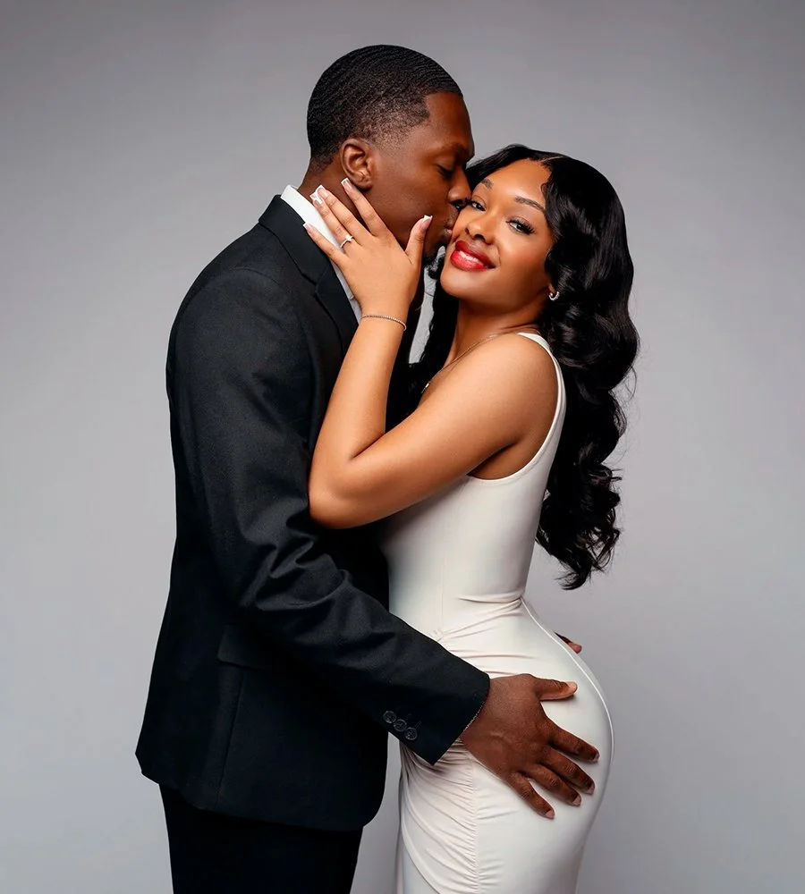 Elegant couple portrait of a man in a black suit kissing his partner in a white fitted dress during a studio engagement photoshoot.