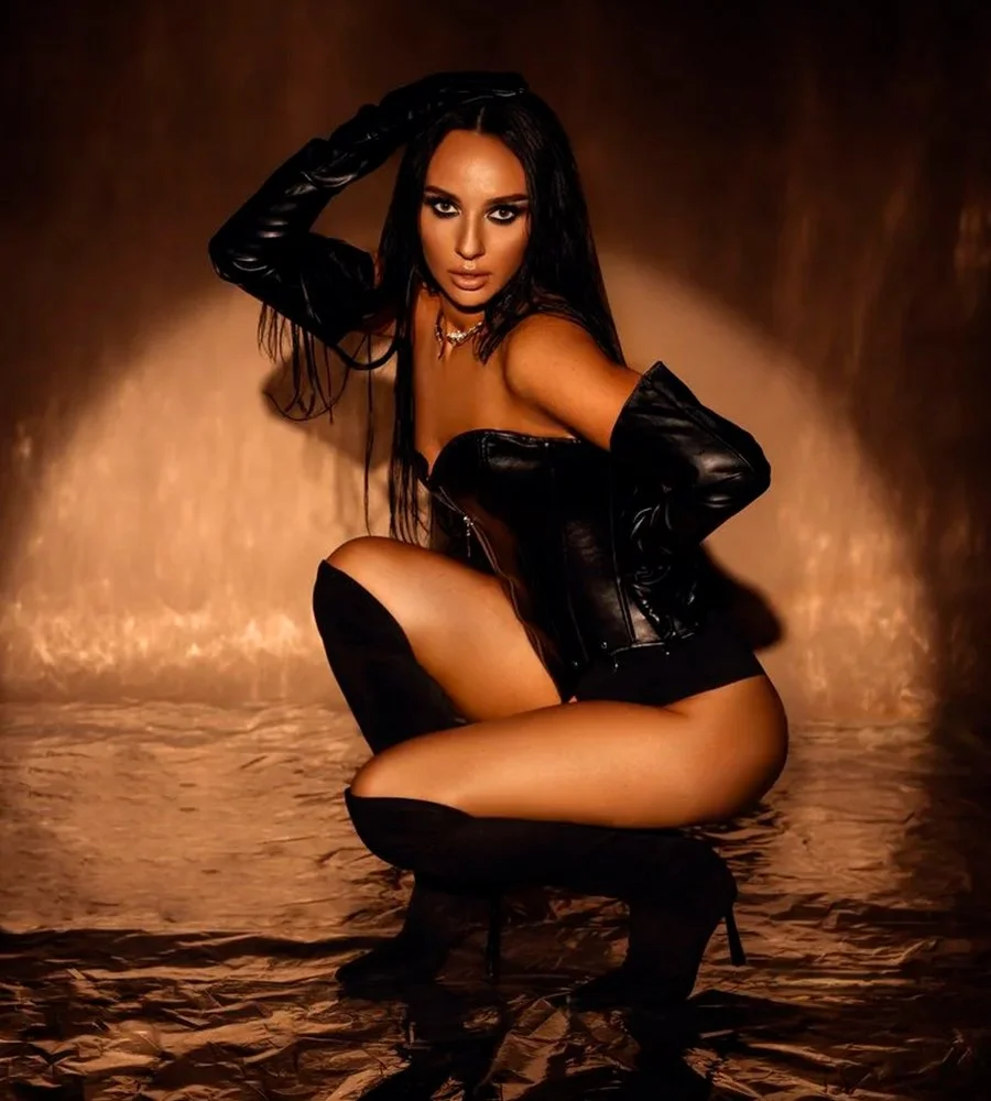 Glamour fashion model in a black leather corset, gloves, and high boots posing in a warm spotlight during a studio photoshoot.